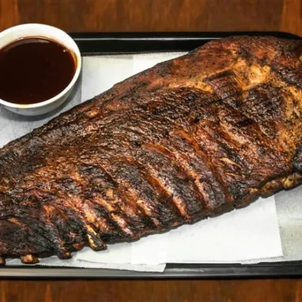 A full rack of Dad's BBQ ribs, dark and smoky, with a side of barbecue sauce.